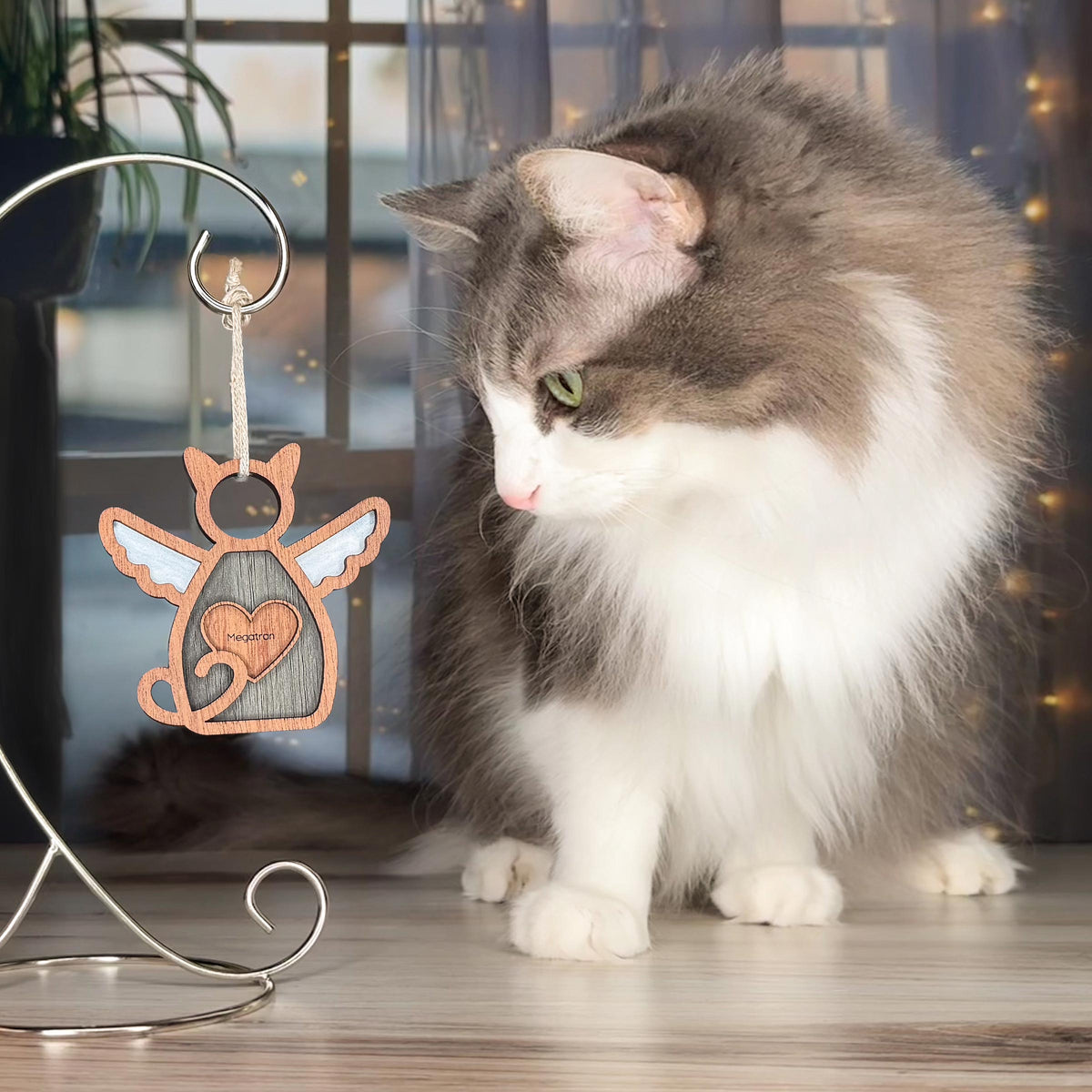 A white and gray cat sits on a wooden floor, gazing at the Forever Loved Cat Angel Ornament for Charity from Forged Flare®, shaped like a winged cat with a heart and hanging from a metal stand. Warm, blurred lights glow behind.