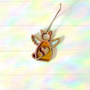 The Forged Flare® Forever Loved Cat Angel Ornament for Charity, featuring wings and a heart cutout, hangs on twine against a pastel rainbow wood background—a heartfelt cat memorial or personalized pet keepsake.