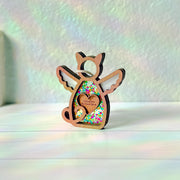 The Forged Flare® Forever Loved Cat Angel Ornament for Charity features a wooden angel with wings and heart cutout, sparkling glitter, and a “Cinnamon, My Sweet Spice” label. It shines on a white surface with soft rainbow reflections.