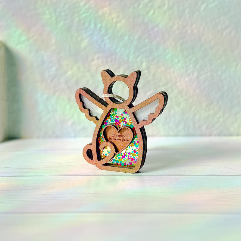 The Forged Flare® Forever Loved Cat Angel Ornament for Charity features a wooden angel with wings and heart cutout, sparkling glitter, and a “Cinnamon, My Sweet Spice” label. It shines on a white surface with soft rainbow reflections.