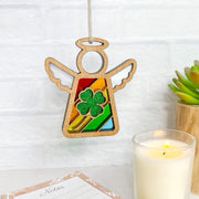 A Forged Flare® Mother's Angels® 3.5" wooden angel figurine with rainbow stripes and a glittery four-leaf clover ornament hangs by a string, displayed with a candle, succulent, and notepad against a white brick wall.