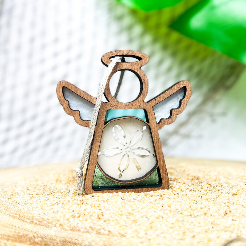 The Sand Dollar Ornament with Imperfections by Forged Flare® is a 3.5" Mother's Angels® handcrafted angel figurine, featuring silver wings, a necklace chain, and sand dollar centerpiece, standing on sand against a bright green backdrop.