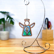 The Forged Flare® Mother's Angels® 3.5" Calla Lily Ornament features stained glass-style wings and hangs on a silver stand atop a wooden surface, with blurred plants and desk items in the background.
