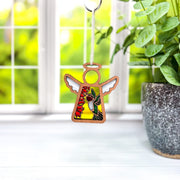 A Forged Flare® Warehouse Return – Ruby-Throated Hummingbird 3.5" Mother’s Angels® ornament hangs in sunlight before a window, while a green potted plant in a gray pot rests on the right side of the white windowsill.