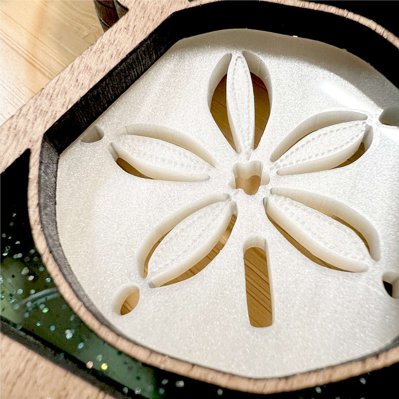 A close-up photo of the Forged Flare® Sand Dollar Ornament with Imperfections—a 3.5" (H) Mother's Angels® figurine—showcasing a handcrafted resin centerpiece, wooden frame, and clear green resin with decorative flecks.