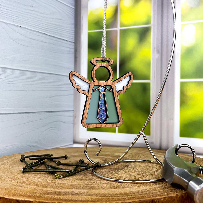 The Forever Your Father Ornament by Forged Flare® is a 3.5" wooden angel figurine with white wings and a blue tie, perfect for Father's Day, displayed on a metal stand with nails, hammer, and greenery visible through a nearby window.