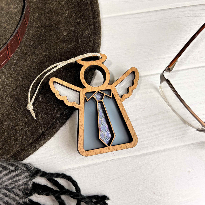 The Forged Flare® Forever Your Father Ornament—a 3.5" Mother's Angels® wooden angel figurine with necktie design—rests on a white surface beside a felt hat, eyeglasses, and scarf; perfect for Father's Day or Christmas.