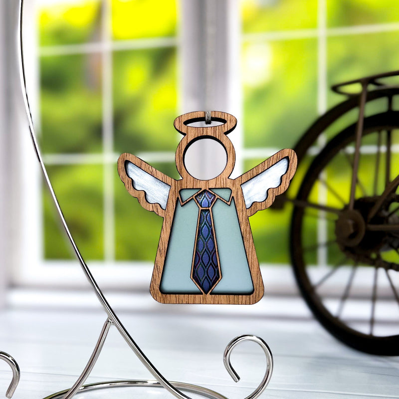 The Forged Flare® Forever Your Father Ornament, a 3.5" Mother's Angels® figurine with white wings and blue tie, hangs on a metal stand—ideal for Father's Day. Green foliage and a bicycle wheel appear through the window behind it.