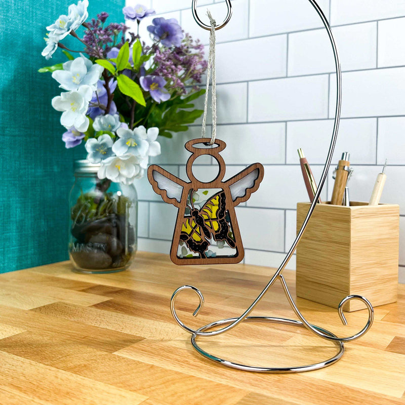 A Warehouse Return - Swallowtail Butterfly Ornament (3.5" H) from Mother's Angels® by Forged Flare® hangs on a metal stand atop a wooden counter, with flowers in a jar and a wooden pen holder against white subway tiles in the background.