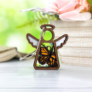 The Monarch Butterfly Ornament with Imperfections, a 3.5" Mother's Angels® angel figurine by Forged Flare®, rests on a white surface near books and pink flowers—an ideal memorial gift to cherish special memories.
