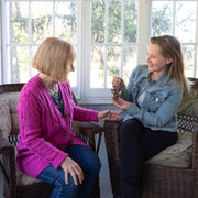 Two women sit on wicker chairs by a sunlit window. Smiling, the younger hands a key to the older—perhaps celebrating finding a Warehouse Return - Sunflower Ornament, a 3.5" Mother's Angels® Angel Figurine by Forged Flare®.