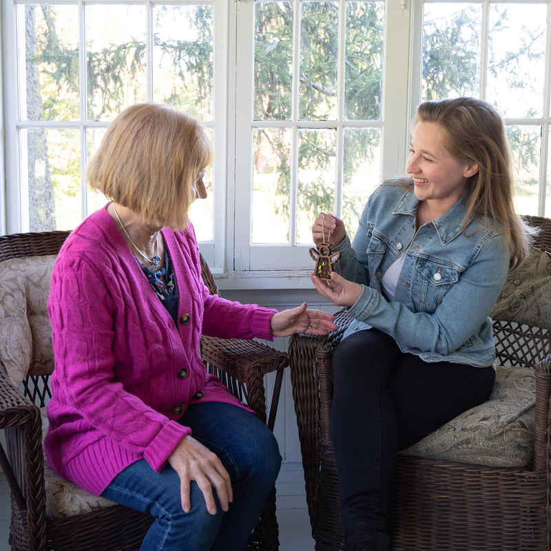 Two women sit on wicker chairs by a sunlit window. Smiling, the younger hands a key to the older—perhaps celebrating finding a Warehouse Return - Sunflower Ornament, a 3.5" Mother's Angels® Angel Figurine by Forged Flare®.