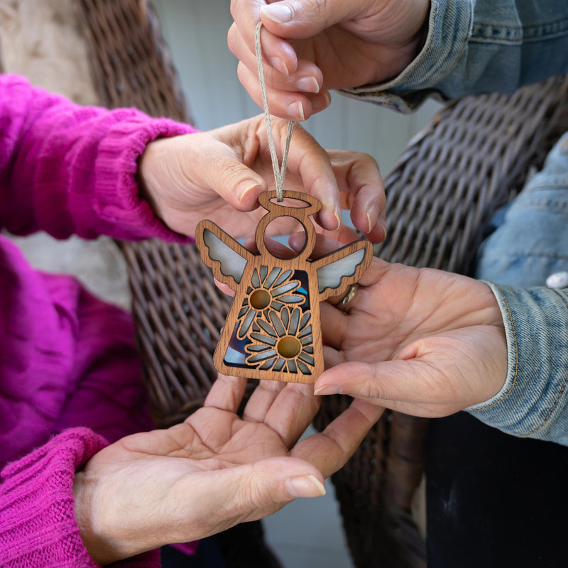 Three hands—one in a pink sweater, one in a denim jacket, and one bare—hold the Forged Flare® Warehouse Return - Daisy Ornament, a 3.5" Mother's Angels® angel figurine with painted flowers and wings. Great for gifting or rediscovery from Amazon warehouse returns.