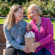 A young and an older woman sit on a bench outdoors, smiling as the older woman holds a Forged Flare® Warehouse Return - Sunflower Ornament 3.5" (H) Angel Figurine | Mother's Angels®, while the younger woman reaches for it, both appearing joyful.