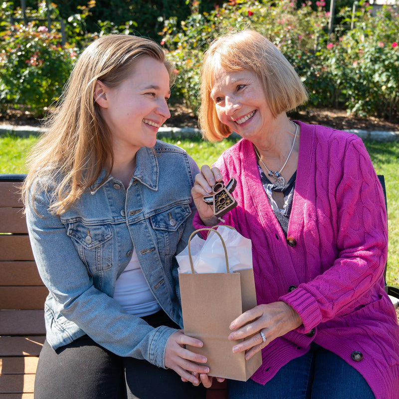 A young and an older woman sit on a bench outdoors, smiling as the older woman holds a Forged Flare® Warehouse Return - Sunflower Ornament 3.5" (H) Angel Figurine | Mother's Angels®, while the younger woman reaches for it, both appearing joyful.