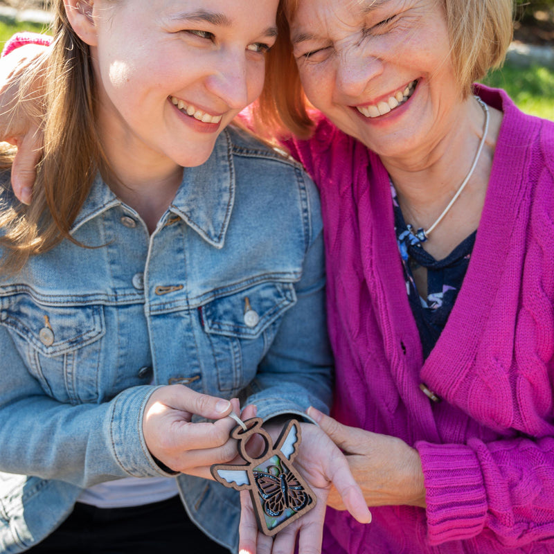 Two women, one in a denim jacket and the other in a pink cardigan, smile as they hold the Forged Flare® Mother's Angels® Monarch Butterfly Ornament with Imperfections—an angel figurine that brings meaning and connection to their moment outdoors.