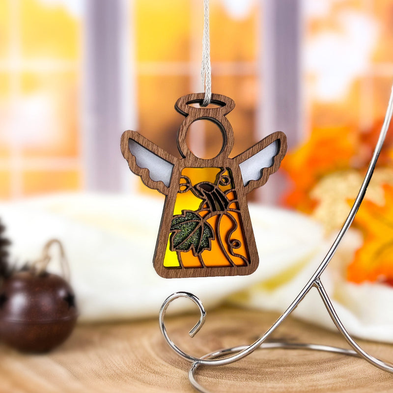 A charming Festive Pumpkin Ornament from Forged Flare® hangs from a white string, featuring a 3.5" wooden angel figurine with an orange body adorned with an embossed leaf and acorn design. Set against a blurred, autumn-themed background filled with warm tones and foliage, this delightful piece from the Mother's Angels® collection makes for a perfect fall decoration or thoughtful memorial gift.