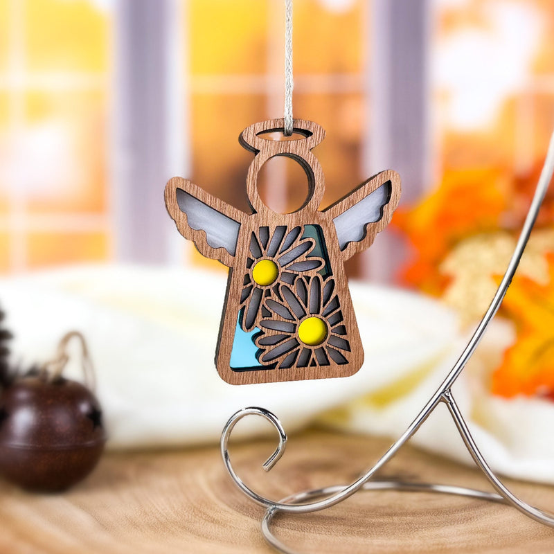 A 3.5" Mother’s Angels® Daisy Ornament by Forged Flare®, featuring cut-out wings and two daisies, hangs from a string. The softly blurred background shows a window and autumn decor, as displayed on Amazon.