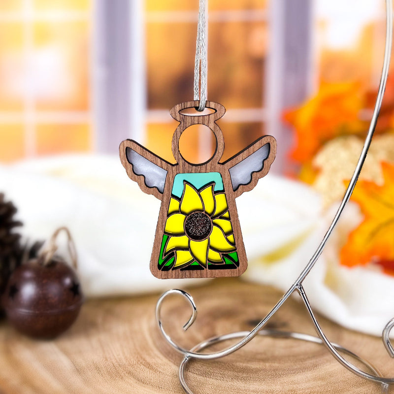 The Forged Flare® Mother's Angels® Sunflower Ornament, a 3.5" wooden angel figurine with stained glass-style wings, hangs on a stand amid softly blurred autumn leaves and a pinecone. This is a Warehouse Return item.