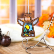 The Warm Welcome Ornament with Imperfections by Forged Flare® is a 3.5" handcrafted Mother's Angels® wooden angel figurine, featuring cutout trees, a log cabin, and yellow moon, displayed on a metal stand against an autumn-hued window.