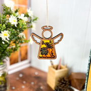 The Forged Flare® Festive Pumpkin Ornament, a 3.5" Mother's Angels® angel figurine, hangs indoors by a string as cozy Thanksgiving decor amid jarred flowers, books, and a softly blurred window.