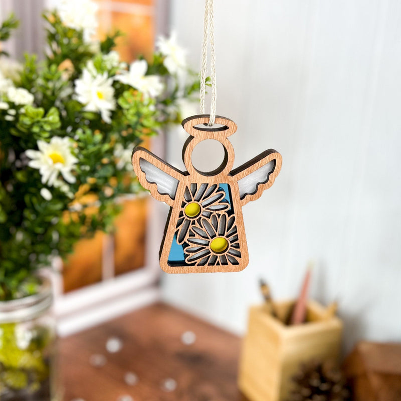 A Forged Flare® Mother's Angels® Daisy Ornament—3.5" (H) angel figurine with daisy flower cutouts and yellow centers—hangs before a blurred background featuring white flowers, a window, and a wooden pen holder.