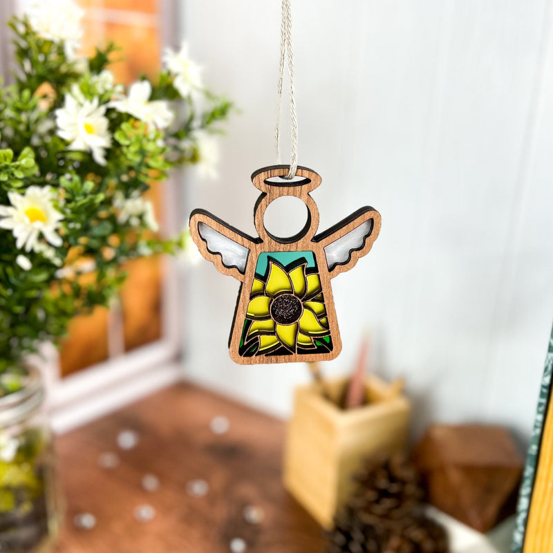 A Forged Flare® Mother's Angels® 3.5" sunflower ornament from a Warehouse Return hangs on a string, with white daisies, a window, and a small wooden box in the background.