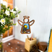 The Warm Welcome Ornament by Forged Flare® is a 3.5" angel figurine from Mother's Angels®, featuring trees, mountains, and a moon cutout, displayed near a window with white flowers and books on a wooden table.