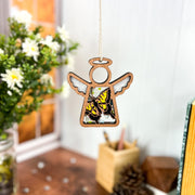 A Forged Flare® Mother’s Angels® 3.5" Swallowtail Butterfly Ornament hangs in a cozy room filled with flowers, books, and art supplies on a wooden desk, sunlight streaming through the window behind it.