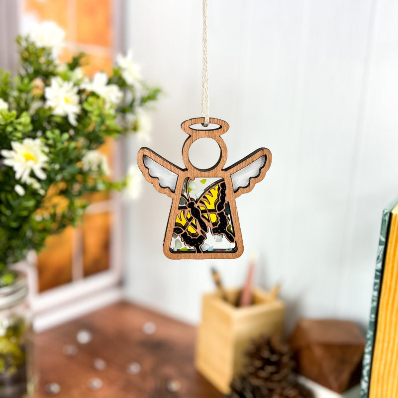 A Forged Flare® Mother’s Angels® 3.5" Swallowtail Butterfly Ornament hangs in a cozy room filled with flowers, books, and art supplies on a wooden desk, sunlight streaming through the window behind it.