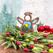 The Forged Flare® Warm Welcome Ornament, a 3.5" Mother's Angels® angel figurine, rests on green foliage and red berries, showcasing painted trees, a cozy cabin, and yellow moon against a softly blurred backdrop.