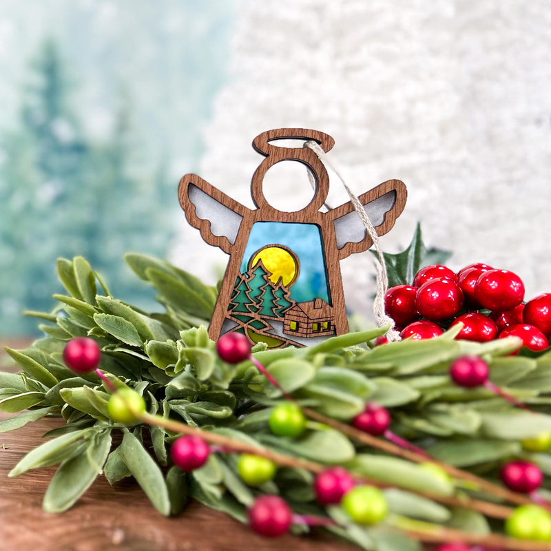 The Forged Flare® Warm Welcome Ornament, a 3.5" Mother's Angels® angel figurine, rests on green foliage and red berries, showcasing painted trees, a cozy cabin, and yellow moon against a softly blurred backdrop.