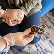 Two people sit together; one holds a Warehouse Return - Sunflower Ornament 3.5" Angel Figurine by Forged Flare®, while the other shows rings and a smartwatch. A patterned rug and blue couch are visible.