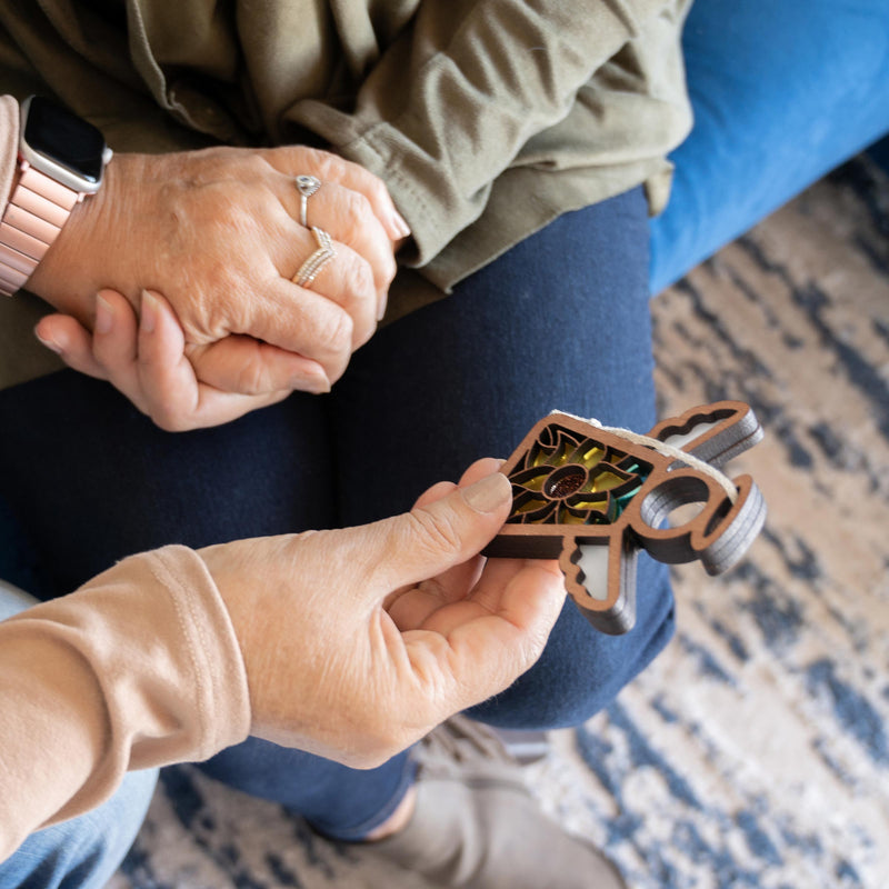 Two people sit together; one holds a Warehouse Return - Sunflower Ornament 3.5" Angel Figurine by Forged Flare®, while the other shows rings and a smartwatch. A patterned rug and blue couch are visible.