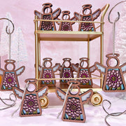 Forged Flare® Mother's Angels® Chrysanthemum Ornament with Imperfections features a 3.5" angel figurine with purple flowers and white wings, displayed on a gold stand with a chrysanthemum centerpiece against a soft pink background and white trees.