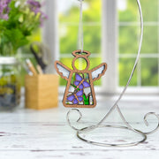 A Warehouse Return Playful Purple Butterflies Angel Ornament by Forged Flare® (Mother’s Angels®, 3.5" H) hangs on a silver stand, with a bright window, green plants, and art supplies on a white distressed surface in the background.