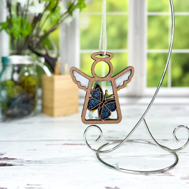 The Forged Flare® Warehouse Return - Morpho Blue Butterfly Ornament is a 3.5" Mother's Angels® figurine with white wings and a blue glitter butterfly, hanging on a metal stand. Blurred background features window, greenery, and jar on white table.