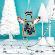 A 3.5" Morpho Blue Butterfly Angel Figurine by Forged Flare® Mother's Angels®, available as a warehouse return, features a blue glitter butterfly design and hangs on a silver stand against a bright turquoise backdrop with white Christmas trees and gems.