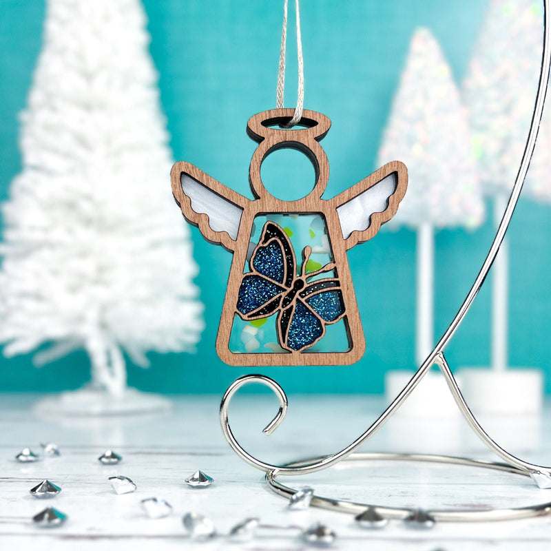 A 3.5" Morpho Blue Butterfly Angel Figurine by Forged Flare® Mother's Angels®, available as a warehouse return, features a blue glitter butterfly design and hangs on a silver stand against a bright turquoise backdrop with white Christmas trees and gems.