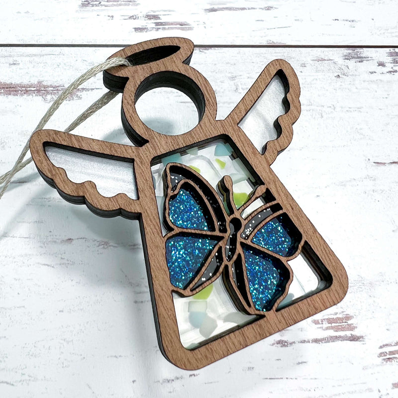 The Forged Flare® Mother's Angels® Warehouse Return Morpho Blue Butterfly Ornament is a 3.5" wooden angel figurine with a blue glittery butterfly and hanging string, shown on a white wooden surface.