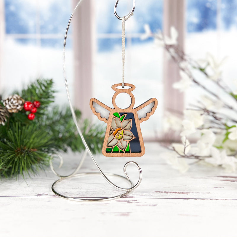 A Narcissus Ornament from Forged Flare's Mother's Angels® collection hangs gracefully on a silver stand in front of a snowy window. The 3.5" angel figurine features an intricate narcissus flower design at its center. Festive greenery, white flowers, and red berries adorn the background, creating a charming wintry ambiance.