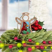 The Forged Flare® Mother's Angels® Winter Cardinal Ornament with Imperfections is a 3.5" handcrafted angel figurine featuring a cardinal centerpiece, holly design, and green wreath with red berries against a snowy window backdrop.