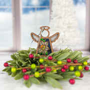 The Forged Flare® Narcissus Ornament is a 3.5" Mother's Angels® wooden angel figurine featuring a painted narcissus, December's birth flower, standing amid green leaves and red berries with a snowy window and white Christmas tree behind.