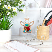 The 3.5" Teacher Ornament with Imperfections by Forged Flare® (Mother’s Angels®), a handcrafted angel figurine featuring an apple and pencil, hangs on a stand, creating a charming teacher-themed centerpiece for any light desk.