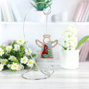 A Forged Flare® Mother's Angels® Winter Cardinal Ornament with Imperfections, a 3.5" angel figurine, hangs from a silver stand on a white table. White flowers, green leaves, and blurred shelves decorate the background.