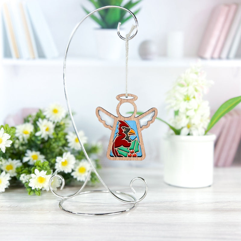 A Forged Flare® Mother's Angels® Winter Cardinal Ornament with Imperfections, a 3.5" angel figurine, hangs from a silver stand on a white table. White flowers, green leaves, and blurred shelves decorate the background.