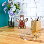 The Gladiolus Ornament, a 3.5" angel figurine from Forged Flare's Mother's Angels®, hangs from a silver stand on a desk, with a jar of purple and white flowers and a wooden holder filled with pens and pencils nearby.