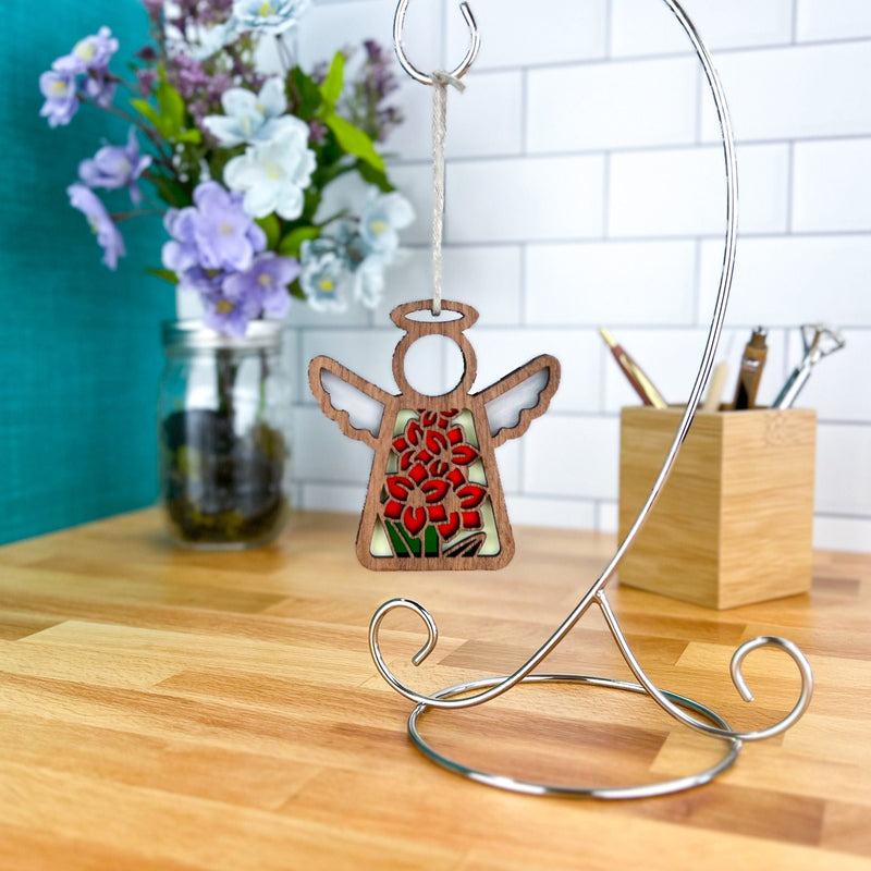 The Gladiolus Ornament, a 3.5" angel figurine from Forged Flare's Mother's Angels®, hangs from a silver stand on a desk, with a jar of purple and white flowers and a wooden holder filled with pens and pencils nearby.