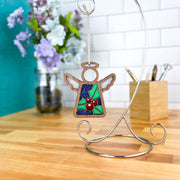 The Forged Flare® Holly Ornament, a 3.5" Mother's Angels® figurine with wings, blue glitter and holly leaves with berries, hangs from a metal stand. Flowers in a jar and a wooden pencil holder appear in the background.