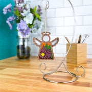 The Forged Flare® Aster Ornament, a 3.5" Mother's Angels® angel figurine featuring the September birth flower, hangs on a metal stand with a jar of flowers and wooden box on a countertop against white tile in the background.